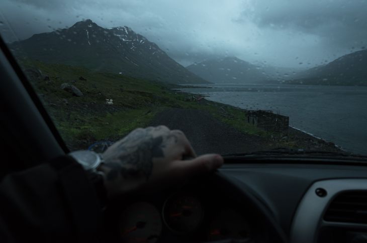 Autoroolist hoiab kinni tätoveeritud mehe käsi. auto esiaknast paistavad mäed ja järv – ilm on vihmane ja sünge.