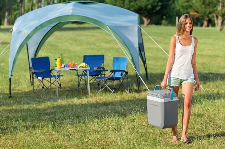 Naine kannab Campingaz Powerbox Plus autokülmikut taustal kokkupandav varjualune piknikulaua ja toolidega.