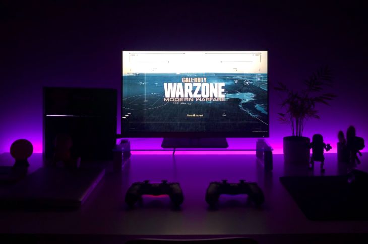 Roosa valgustusega elutuba, diivanilaual on kaks playstation 4 pulti ja teleka ekraanil on Warzone mäng ees.